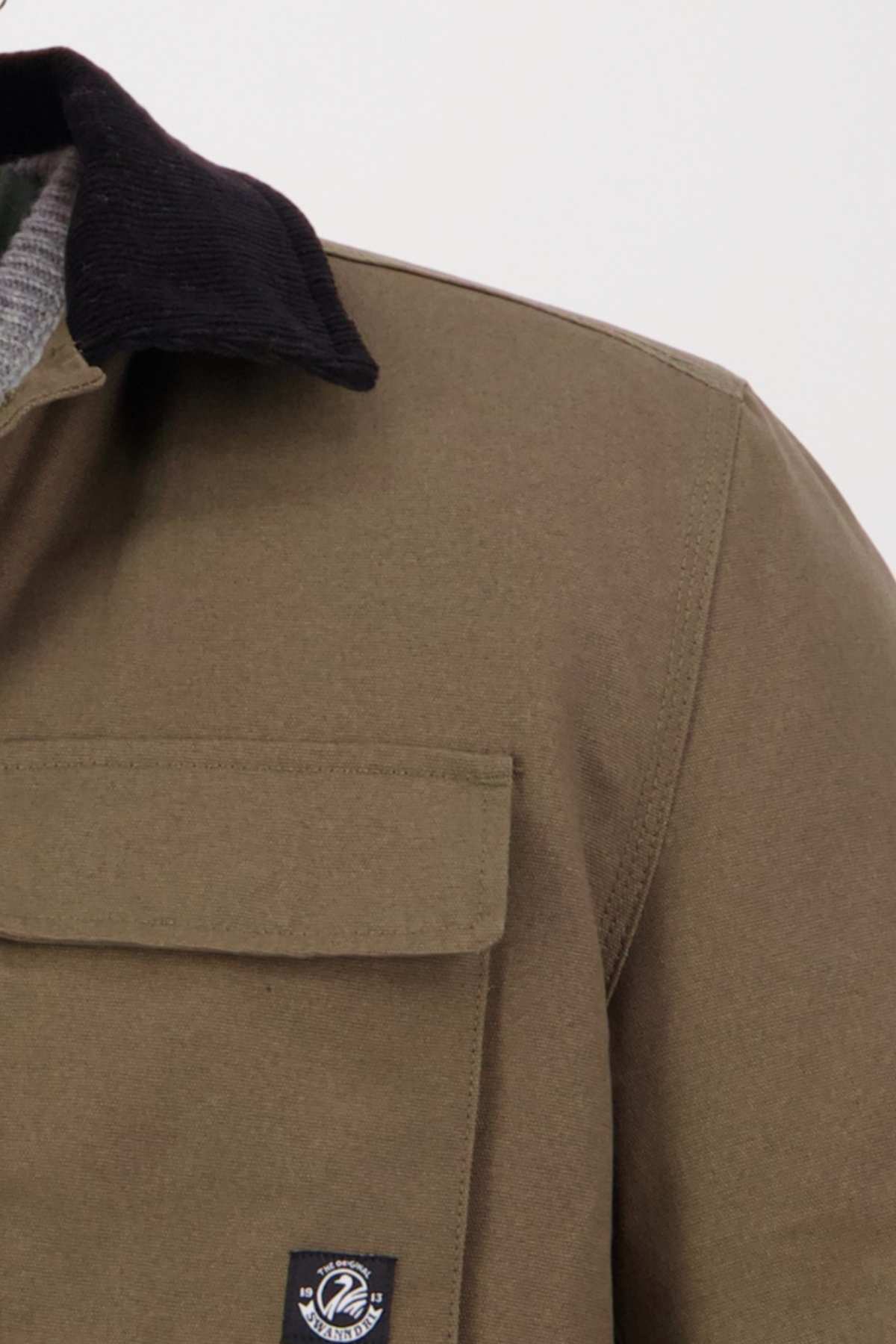Close-up of a person wearing the Swanndri Barrytown v3 Canvas Work Jacket in khaki, featuring a black collar and front pocket with a small logo patch. The collar of a gray sweater or shirt peeks out underneath against a plain white background.