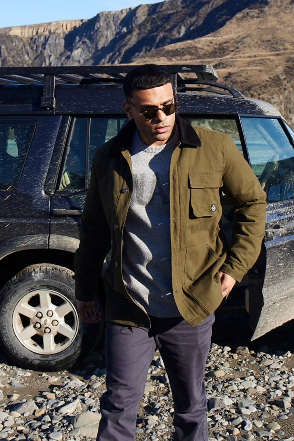 A man wearing a Swanndri Barrytown v3 Canvas Work Jacket, gray sweater, and dark pants stands before a muddy SUV on rocky terrain. The rugged backdrop with rocky hills and blue sky highlights an ideal outdoor setting for testing this durable workwear.