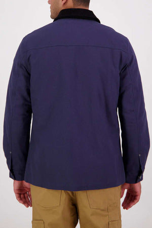 A man stands with his back to the camera, wearing a dark blue Swanndri Barrytown v3 Canvas Work Jacket with a black collar and tan pants. The cotton canvas material contrasts with the plain, light-colored background.