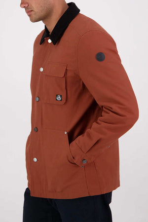 Someone stands against a plain light background wearing the Swanndri Barrytown v3 Canvas Work Jacket in rust, featuring a black collar, front pockets, and reinforced stitching.
