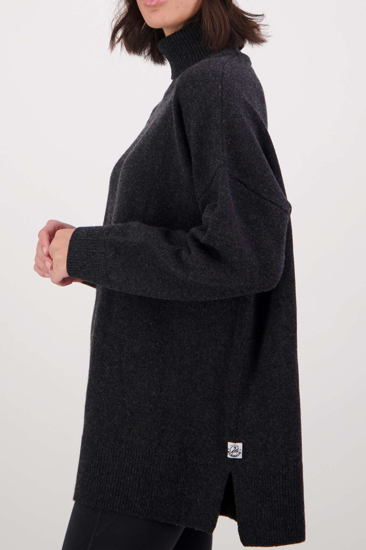 A person in a loose, dark gray Swanndri Brunner Merino Roll Neck Knit sweater with ribbed cuffs and hem, a small patch near the hem, and a side slit stands sideways against a plain light background.