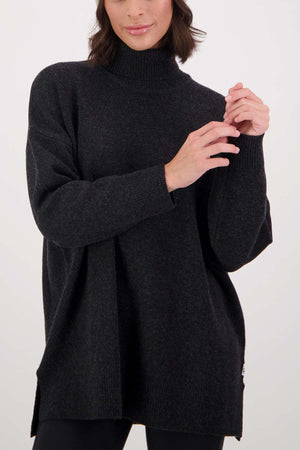 A woman wearing the Swanndri Brunner Merino Roll Neck Knit in a loose, dark fit and black leggings stands against a plain white background, her hands gently clasped near her chest.