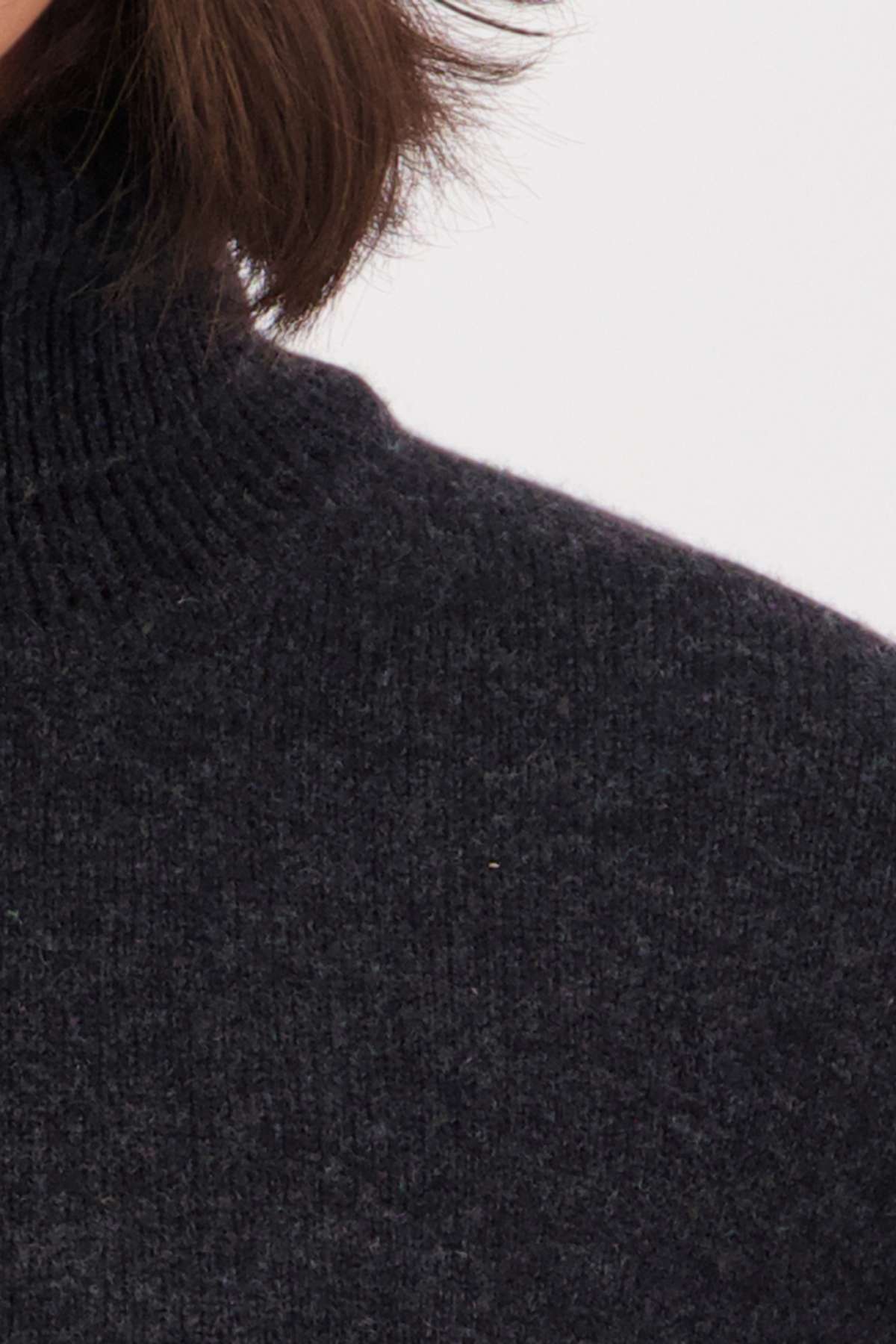Close-up of a person's shoulder and neck wearing the Swanndri Brunner Merino Roll Neck Knit sweater in a dark, textured knit; part of their brown hair is visible against a plain light background.