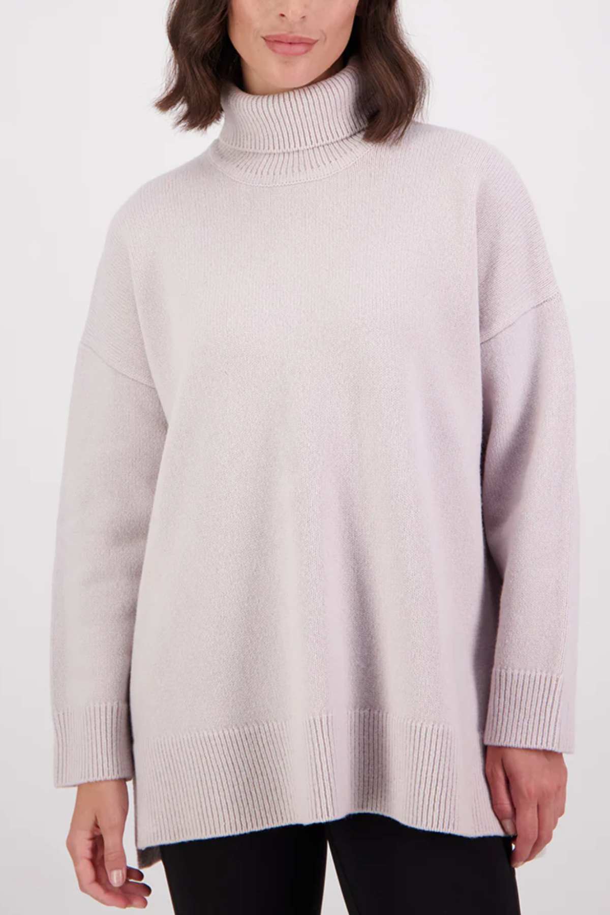 A woman wearing the Swanndri Brunner Merino Roll Neck Knit in light lavender, featuring long sleeves with ribbed cuffs and hem, stands against a plain white background.