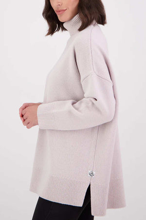 A woman with shoulder-length brown hair wears the Swanndri Brunner Merino Roll Neck Knit in light pink, featuring ribbed cuffs and hem, paired with black pants. She is shown from the side against a plain white background.