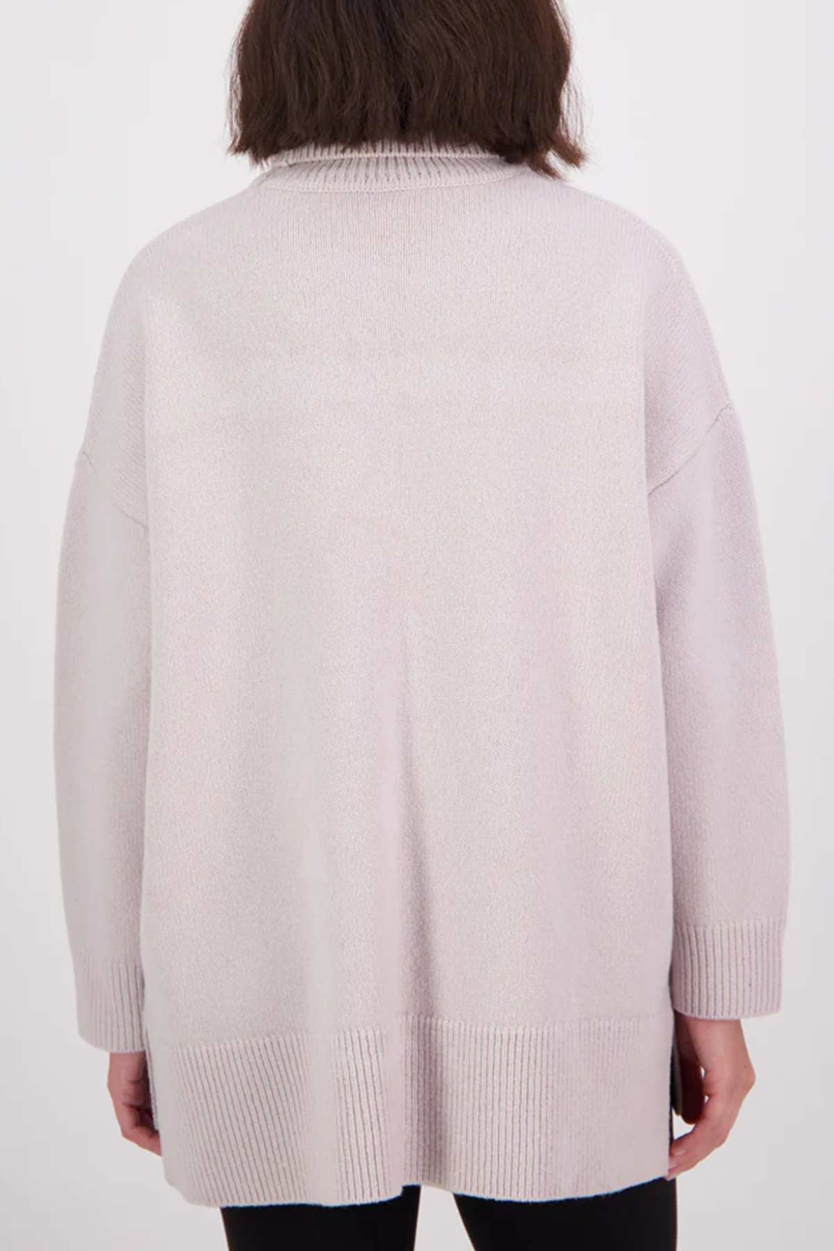 A person with shoulder-length brown hair, seen from the back, wears the Swanndri Brunner Merino Roll Neck Knit in light beige with ribbed cuffs and hem, paired with black pants against a plain white background.