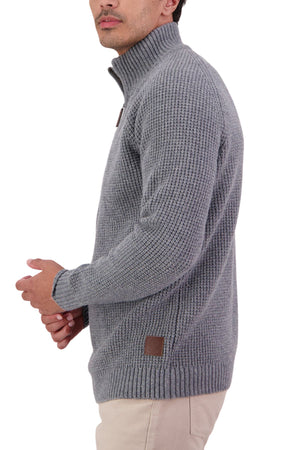 A man wearing the Swanndri Chalky Island Waffle Jumper, a thick light gray sweater with a high collar, and beige pants stands against a white background, facing left.