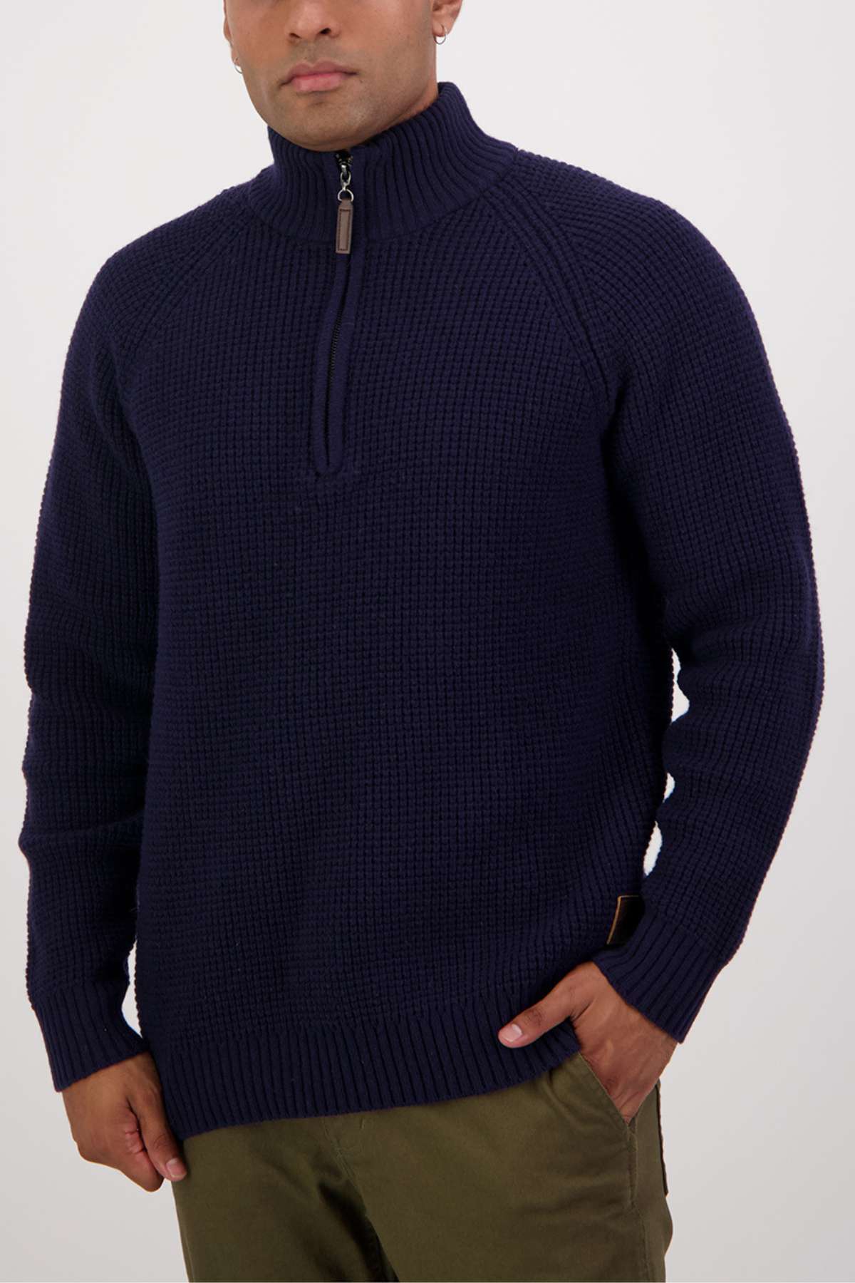 A person is wearing a navy blue Swanndri Chalky Island Waffle Jumper with a cozy knit, half-zip front, and high collar. They have one hand in the pocket of olive green pants and are standing against a plain white background.