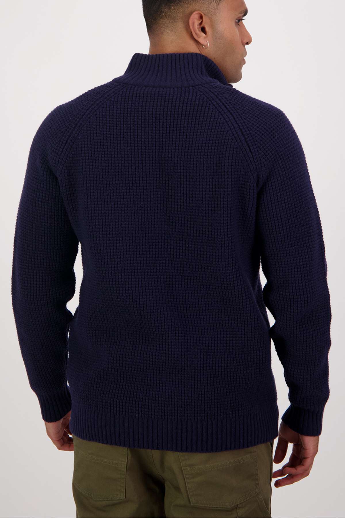 A person with short hair is seen from behind, wearing a Swanndri Chalky Island Waffle Jumper in dark blue. The cozy knit sweater features a high collar and thick material, paired with olive green pants, creating an ensemble that stands out against the plain white background.