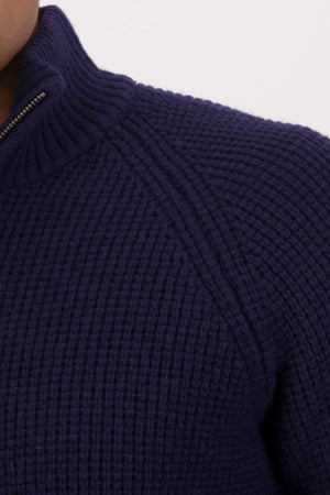 Close-up of a person wearing the Swanndri Chalky Island Waffle Jumper in dark blue, featuring a ribbed high collar and a half zip front. The cozy knit showcases a chunky texture. The person's face is not visible.