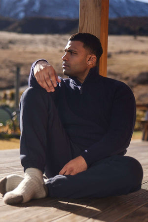 A person sits on a deck, leaning against a wooden post, and gazes into the distance. They are wearing a cozy Swanndri Chalky Island Waffle Jumper with a half zip front, pants, and light-colored socks. The background shows a scenic landscape with mountains and open fields.