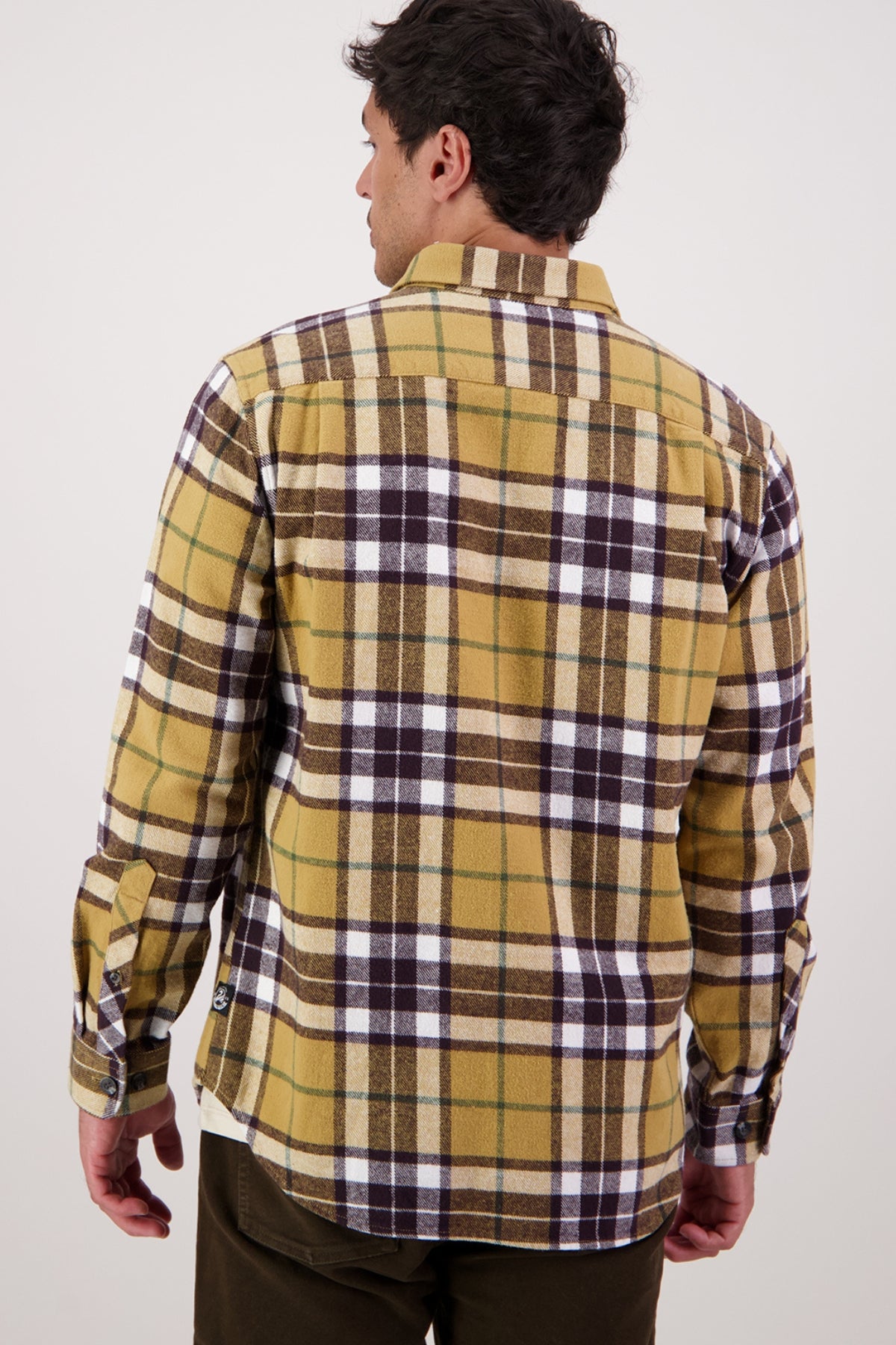 A man is shown from behind wearing the Swanndri Crofton Shirt, featuring yellow and brown plaid in 100% cotton, paired with dark pants and standing against a plain, light background.