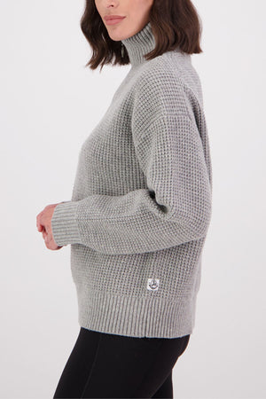 A woman with shoulder-length brown hair models the Swanndri Darlinghurst Zip Jumper in light gray, featuring a ribbed lambswool design and oversized collar, paired with black pants against a white background.