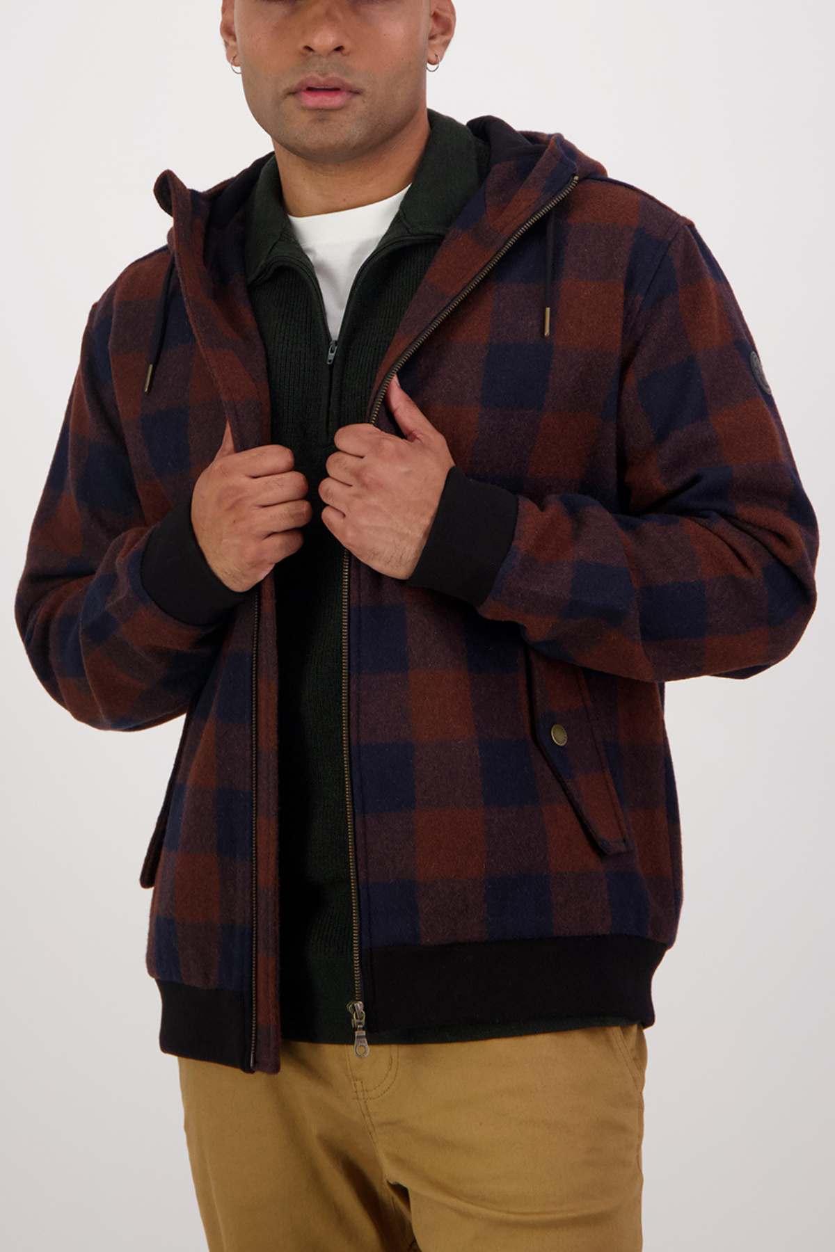 A person wearing a brown and navy checkered jacket over the Swanndri Dunwich Wool Hoody in dark green is adjusting the zipper at mid chest level. They are also wearing tan pants, and the background is plain white.