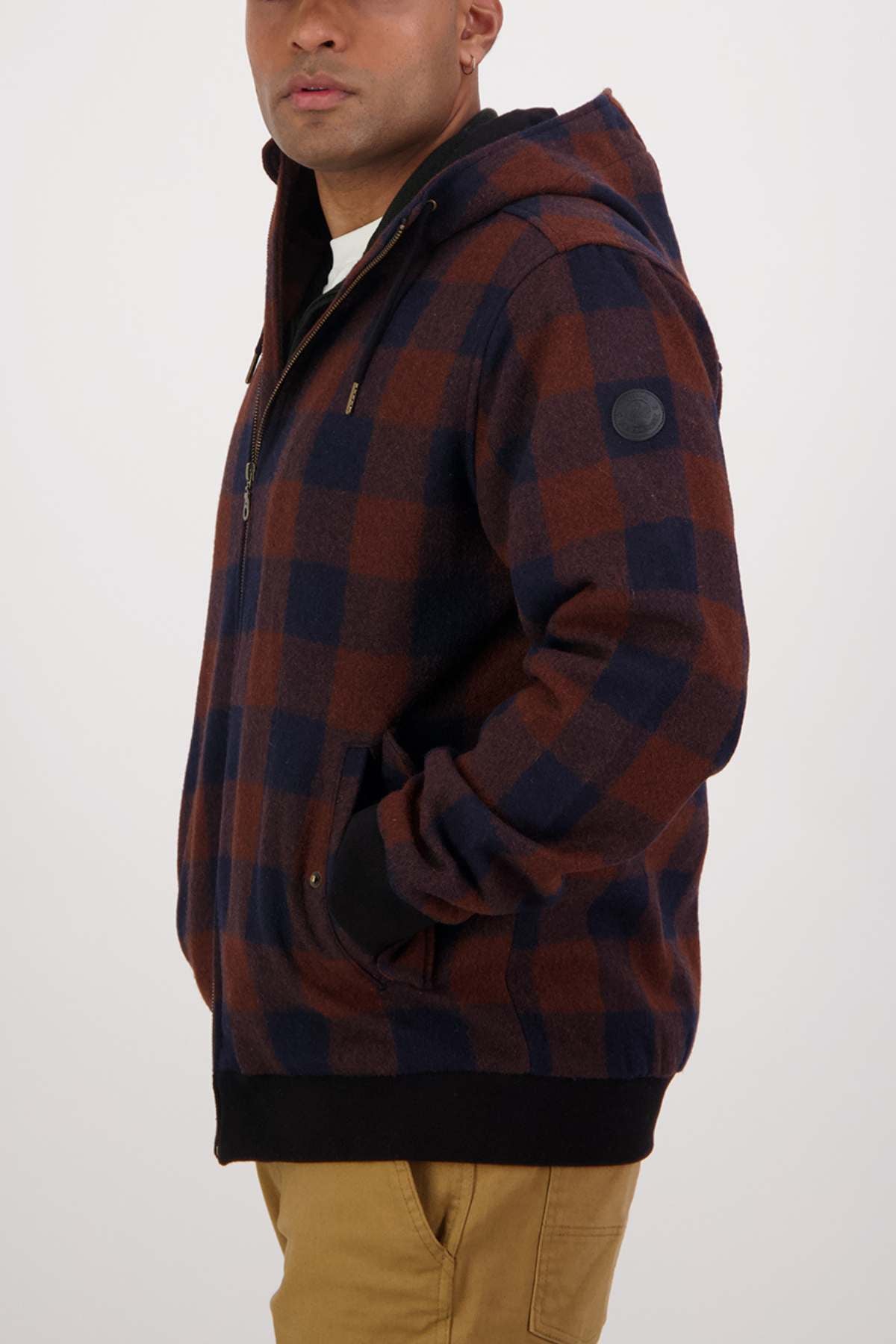 A person wearing a Swanndri Dunwich Wool Hoody in brown and navy checkered merino wool with a hood, standing sideways with hands in their pockets. They have on tan-colored pants. The background is plain white.