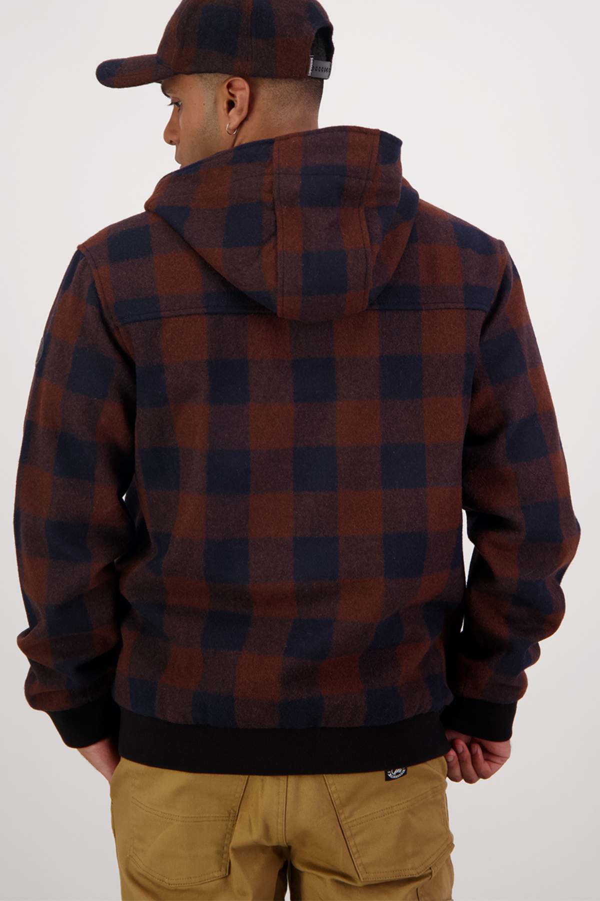 A person wearing a Swanndri Dunwich wool hoody, adorned with a brown and navy checkered pattern, along with a matching cap and beige pants, is facing away from the camera. The hood of the hoody is up, providing extra insulation, and the person has their hands by their sides.