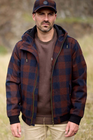 A man wearing a brown and navy plaid jacket over a Swanndri Dunwich Wool Hoody and a matching navy and brown cap stands outdoors in a grassy area with a blurred natural background. He has a beard and a serious expression.