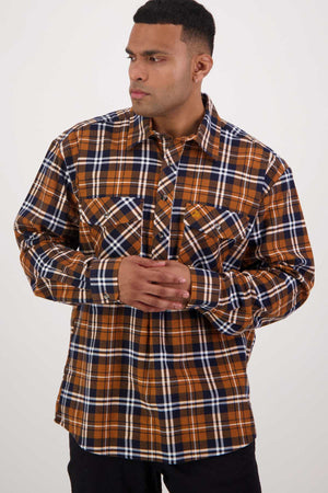 A man stands against a plain background, wearing a long-sleeved, button-up plaid shirt in brown, navy blue, and white brushed cotton from the Swanndri Egmont Flannelette Twin Pack. He has his hands clasped in front of him and is looking to his right with a serious expression.