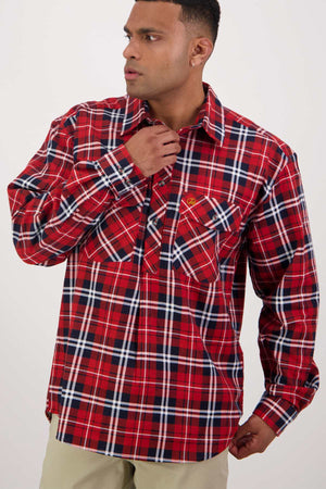 A man wearing a red, white, and black plaid shirt from the Swanndri Egmont Flannelette Twin Pack poses against a neutral background. He holds the collar of his long sleeve shirt with one hand and looks off to the side. He has short hair and is dressed in light-colored pants.