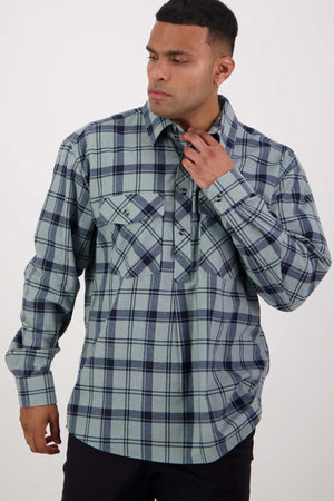 A young man stands against a plain background wearing a long-sleeved, blue and gray plaid flannelette shirt from the Swanndri Egmont Flannelette Twin Pack. He is looking slightly to his right and adjusting the collar of his shirt with his right hand.
