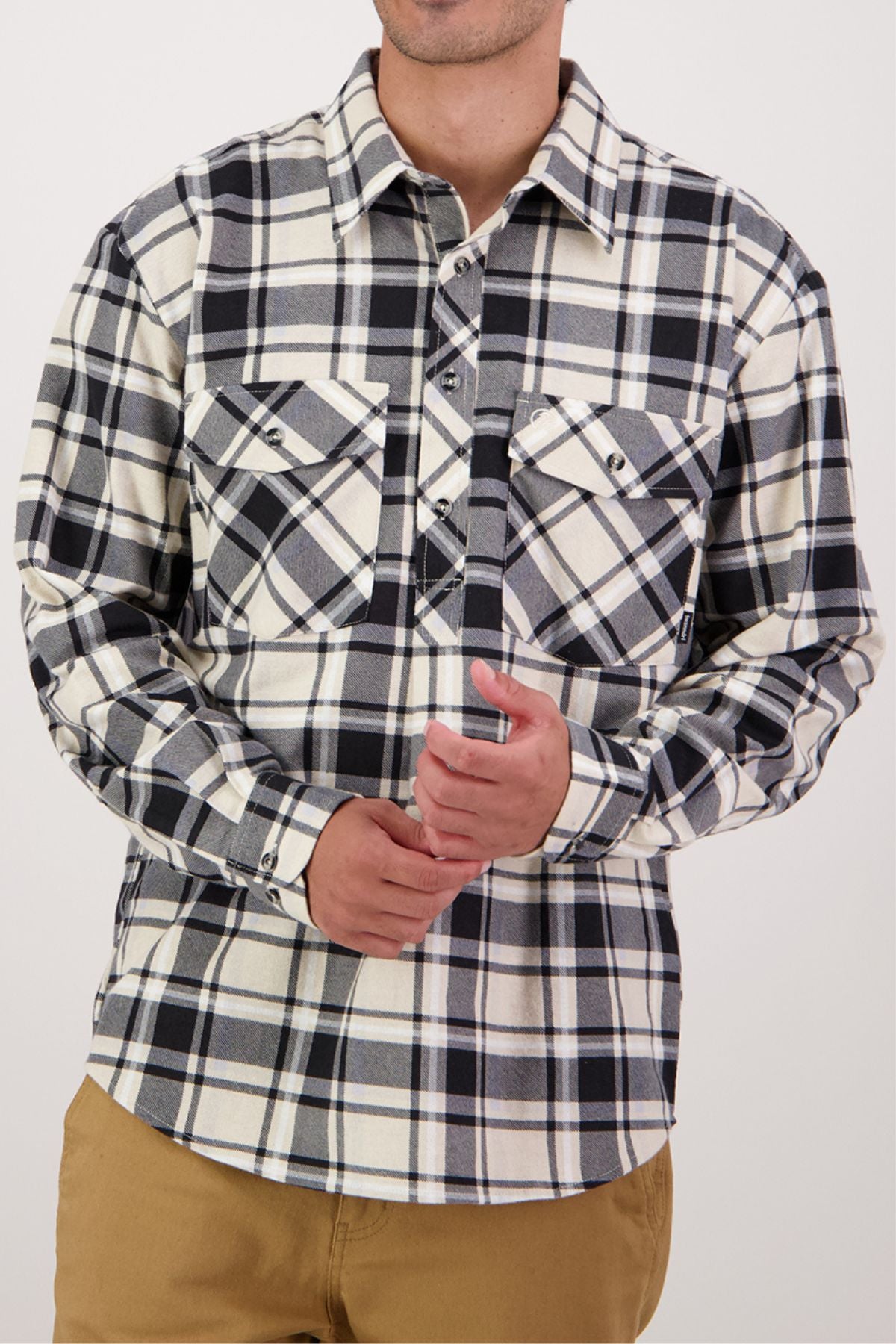 A man stands against a plain light background wearing the Swanndri Egmont Flannelette Twin Pack shirt—black, white, and gray plaid with two chest pockets—and khaki pants. His face is partially out of view.