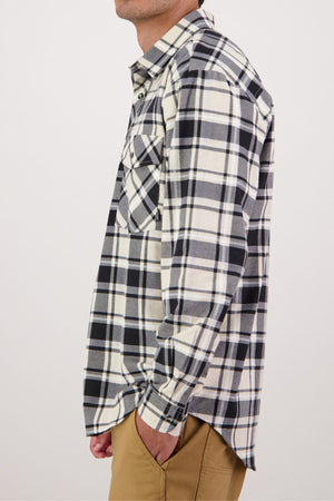 A person wears the Swanndri Egmont Flannelette Twin Pack shirt—black, white, and beige plaid—paired with tan pants. The side view shows one hand in their pocket against a plain white background.