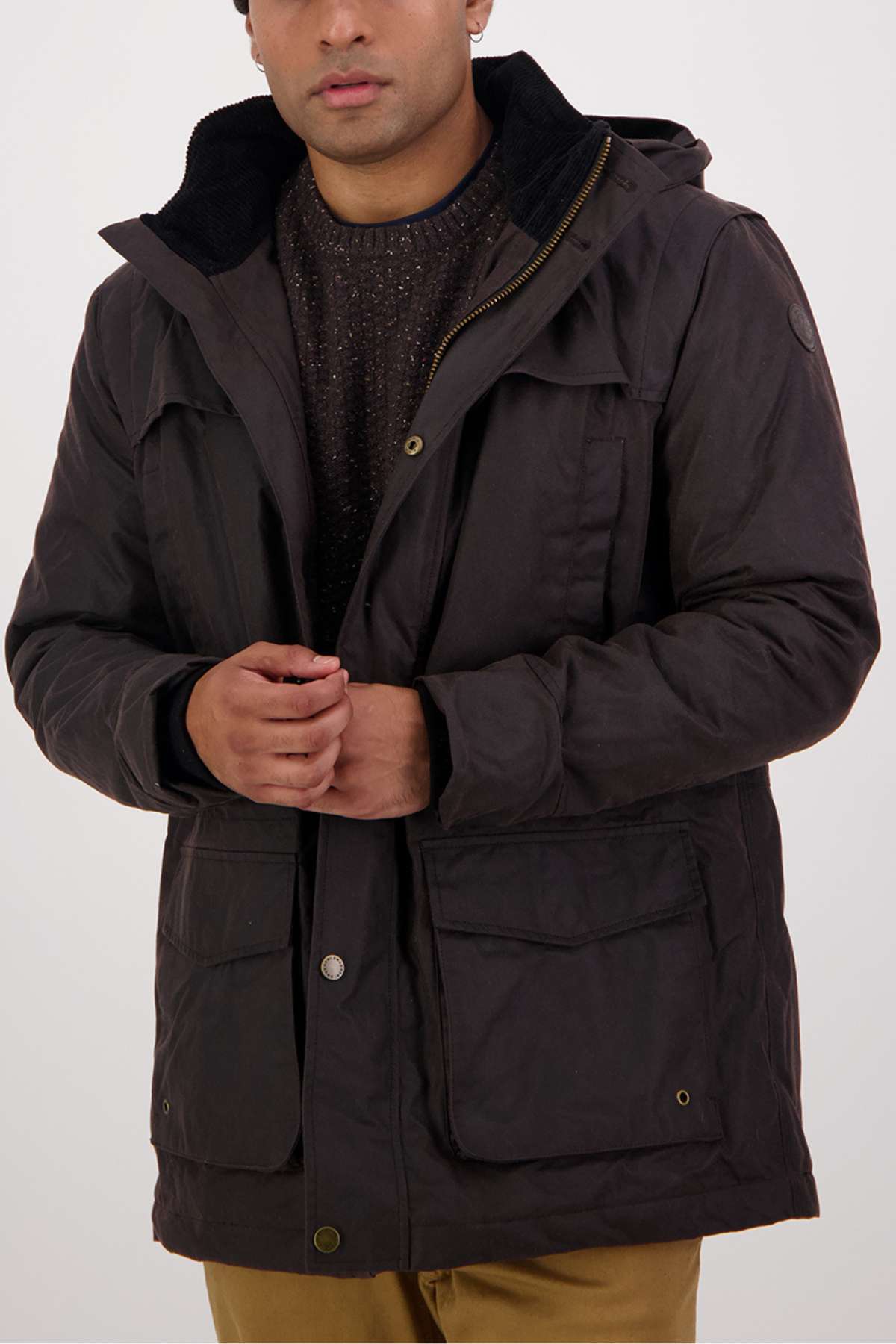 A person wearing a dark brown Swanndri Evans Flat Oilskin Jacket with quilted lining over a textured sweater and tan pants stands against a light background, adjusting the jacket’s front. The person's face is partially visible.