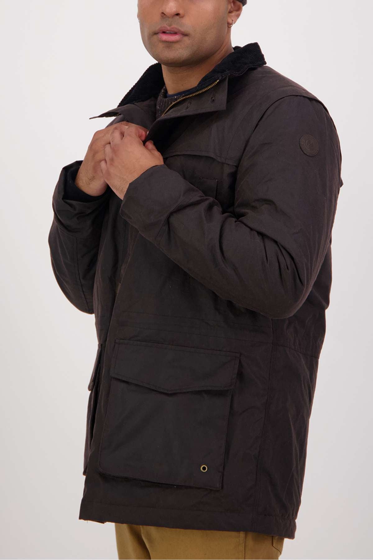 A man wearing a dark brown Swanndri Evans Flat Oilskin Jacket with large front pockets, collar, and quilted lining stands against a plain light background, adjusting his jacket near his chest with his face partially visible.