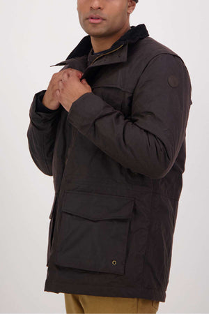 A man wearing a dark brown Swanndri Evans Flat Oilskin Jacket with large front pockets, collar, and quilted lining stands against a plain light background, adjusting his jacket near his chest with his face partially visible.