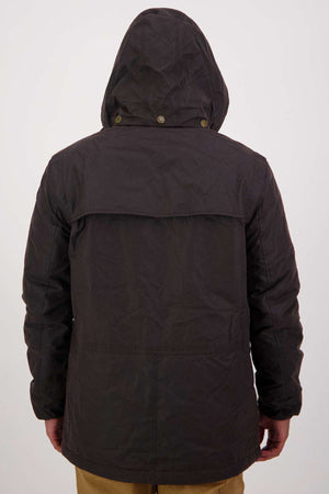 A person is seen from behind wearing a Swanndri Evans Flat Oilskin Jacket with the hood up and brown pants. The jacket has a flap across the upper back and visible seams, standing out against a simple, light background.
