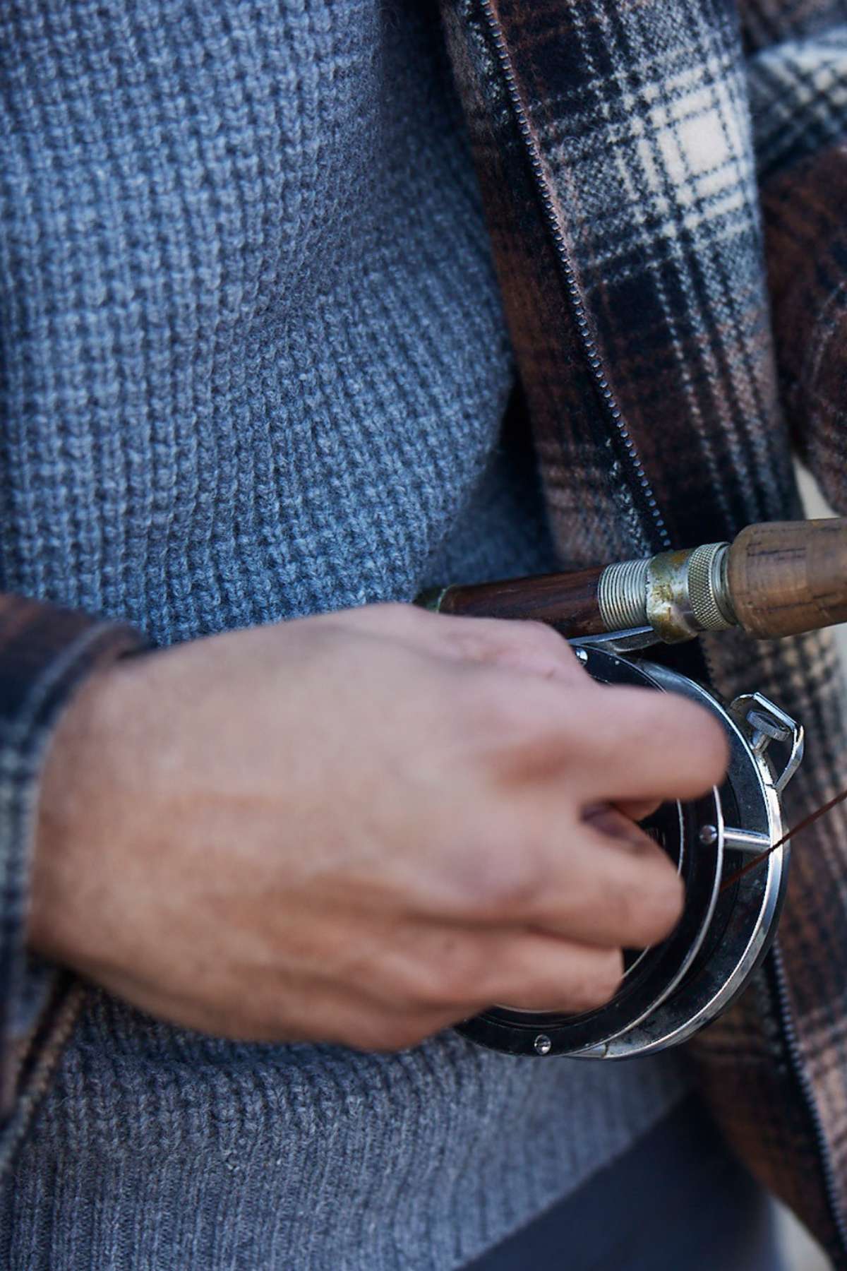 A close-up of someone in a Swanndri Fistral Waffle Knit sweater and plaid jacket holding a fishing rod, adjusting the reel.