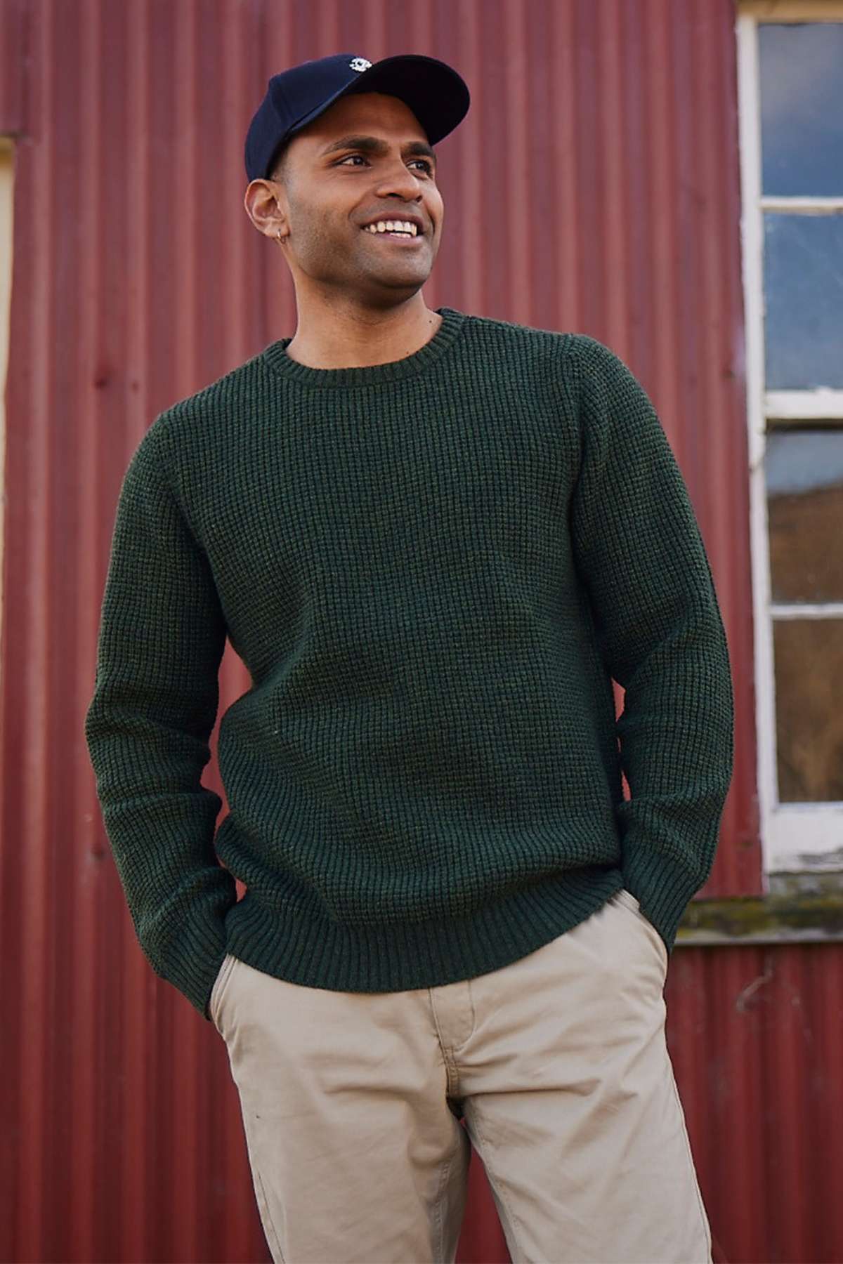 A person in a Swanndri Fistral Waffle Knit sweater in dark green and beige pants smiles while standing in front of a red corrugated metal wall with a window. They are also wearing a dark blue baseball cap.