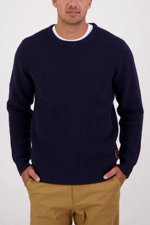 A man wears the Swanndri Fistral Waffle Knit navy sweater, with a subtle waffle pattern, layered over a white t-shirt and tan pants, standing with hands in pockets against a plain white background.