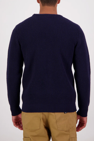 A person faces away from the camera, wearing the Swanndri Fistral Waffle Knit sweater in dark navy and tan pants, set against a plain white background.