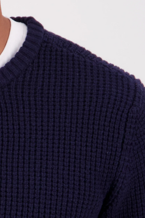 Close-up of the Swanndri Fistral Waffle Knit in navy blue, layered over a white shirt. The image focuses on the textured waffle pattern at the shoulder and neck against a light background.