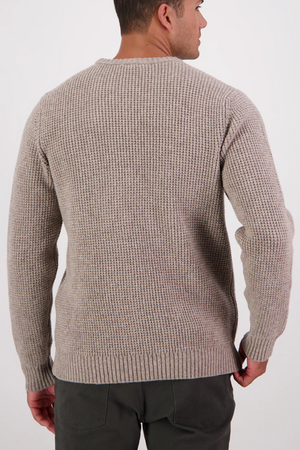 A man is shown from the back wearing the Swanndri Fistral Waffle Knit sweater in light beige, featuring a classic waffle design and ribbed cuffs, paired with dark pants against a plain white background.