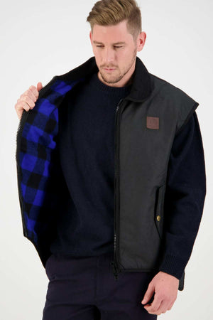 A man in a dark sweater and pants reveals the blue and black plaid interior of his Swanndri Foxton Vest Wool Lined, a sleeveless waterproof black vest made from waxed cotton. He gazes down with a neutral expression against a plain white background.
