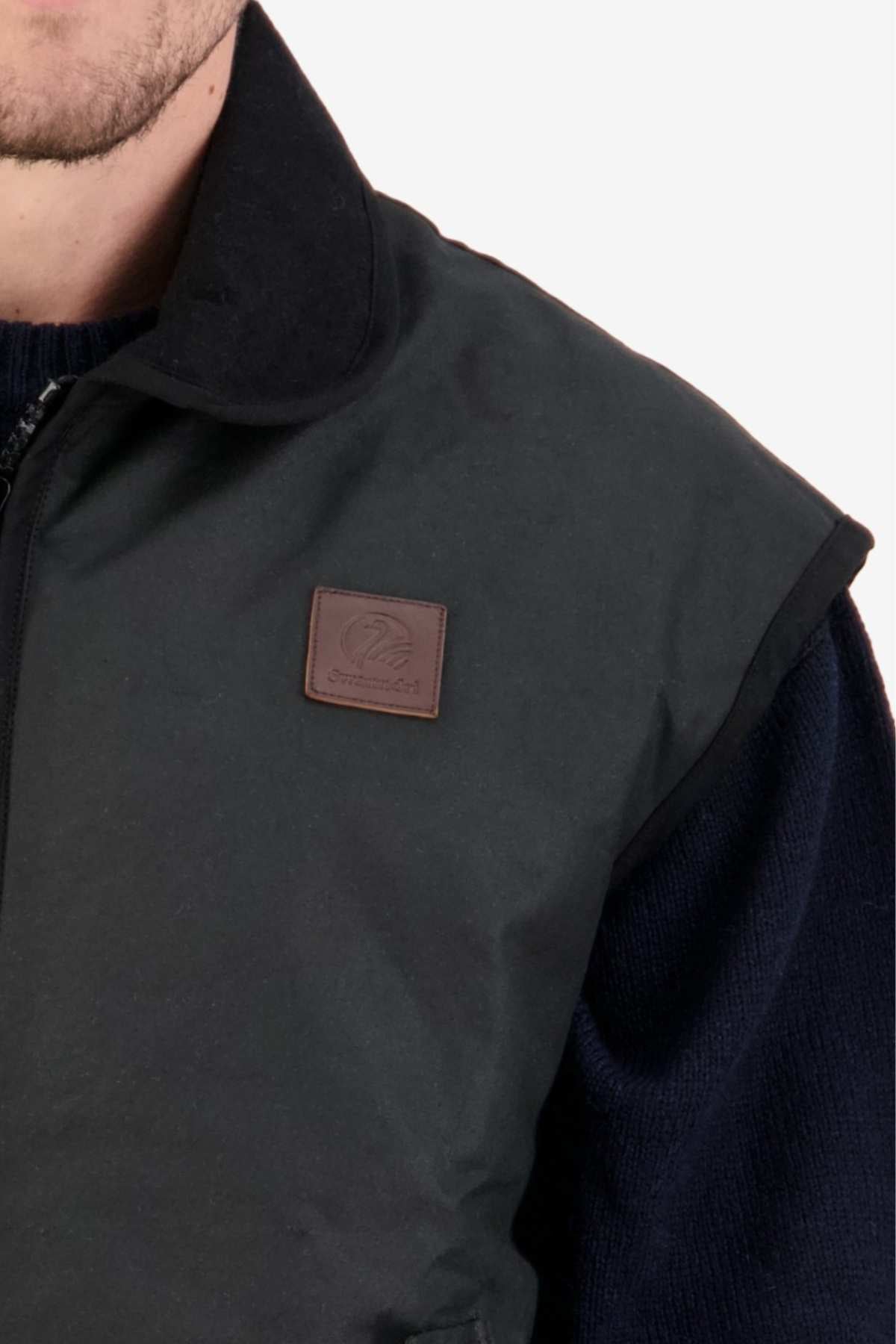Close-up of a person wearing the Swanndri Foxton Vest Wool Lined in dark green with a brown leather patch on the chest. The vest, made of waxed cotton, is layered over a dark sweater, showing the upper torso and part of the person's face.