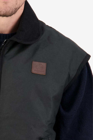 Close-up of a person wearing the Swanndri Foxton Vest Wool Lined in dark green with a brown leather patch on the chest. The vest, made of waxed cotton, is layered over a dark sweater, showing the upper torso and part of the person's face.