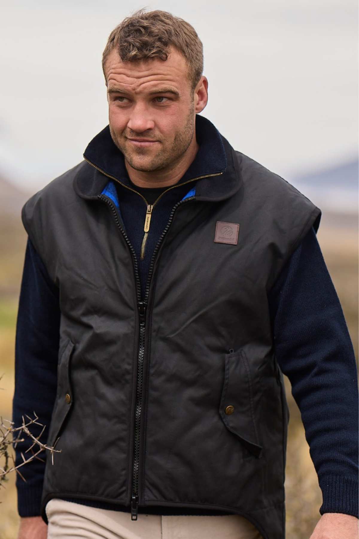 A person in a Swanndri Foxton Vest Wool Lined, black and waterproof with a zipper, stands outdoors wearing it over a dark sweater. They have short hair and are looking slightly to the side against a backdrop of blurred open fields or rural area.