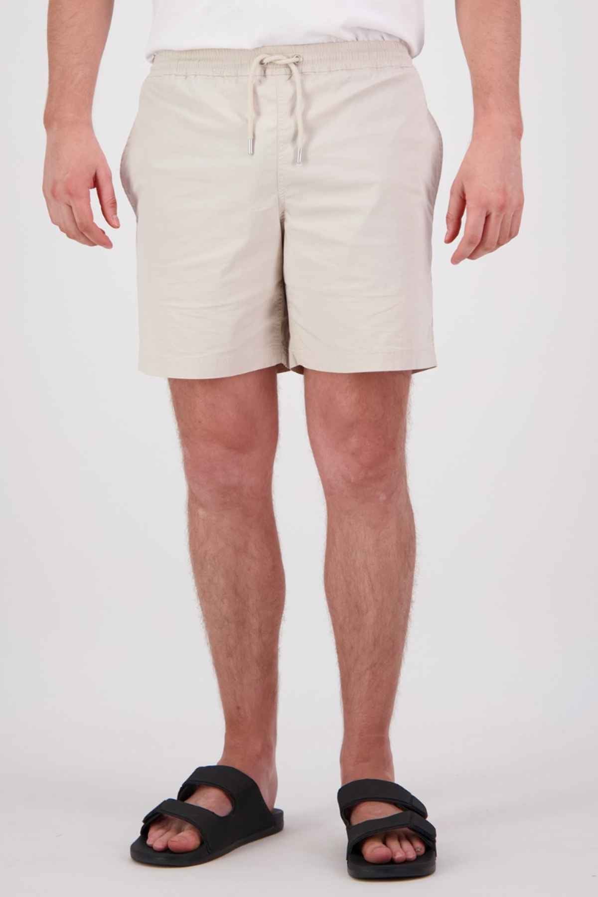 A person wearing Swanndri Frankton Shorts, crafted from a cotton blend, stands against a plain white backdrop. The shorts in light beige boast an elastic waistband with a drawstring and are paired with black sandals. Their hands rest casually at their sides.