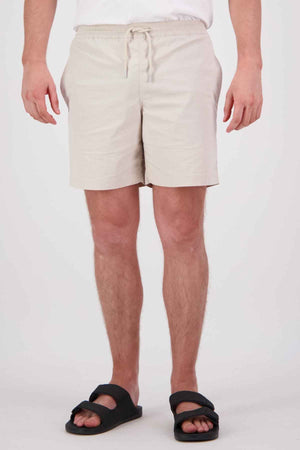 A person wearing Swanndri Frankton Shorts, crafted from a cotton blend, stands against a plain white backdrop. The shorts in light beige boast an elastic waistband with a drawstring and are paired with black sandals. Their hands rest casually at their sides.