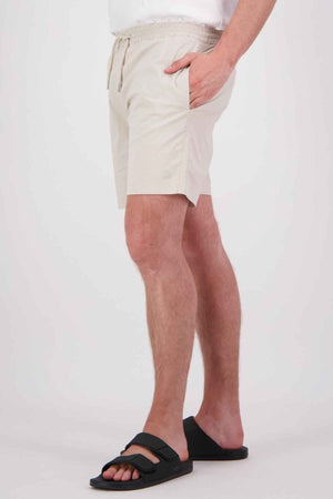 A person wearing the Swanndri Frankton Short, a pair of light beige cotton blend shorts with an elastic waist, hands in pockets, paired with a white shirt and black sandals, against a plain white background.
