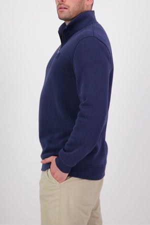 A man stands in profile against a light background, wearing the Swanndri Fundamentals Quarter Zip in navy blue and beige pants, with one hand in his pocket.