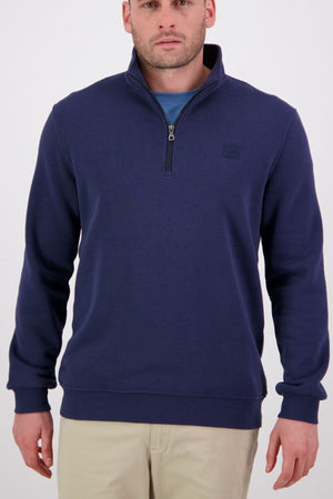 A man wearing a navy blue Swanndri Fundamentals Quarter Zip over a blue shirt and beige pants stands facing forward against a plain white background.