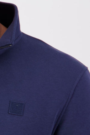 Close-up of a person wearing the Swanndri Fundamentals Quarter Zip in navy blue French Terry, featuring a small square embroidered logo on the chest. Only the shoulder, neck, and part of the collar are visible.
