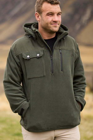 A man is seen standing outdoors wearing the Swanndri High Rock Fleece Pullover, an olive green jacket with multiple pockets and a hood, crafted from waterproof fabric. He is looking slightly to his left with his hands in his pockets. The background features an out-of-focus natural landscape with earthy tones.
