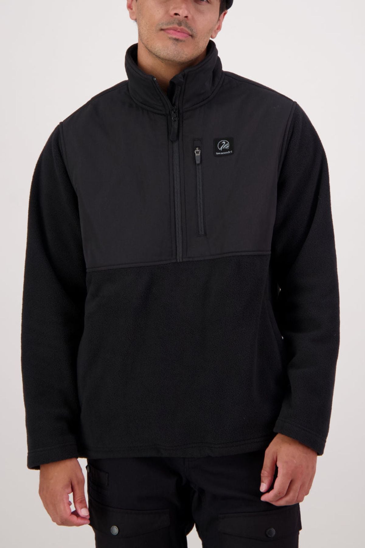A person wears a black Swanndri High Rock Hybrid Fleece Pullover, made from recycled polyester with a long-sleeve quarter-zip, small zippered chest pocket, and logo patch on the left chest. The background is plain and light-colored.