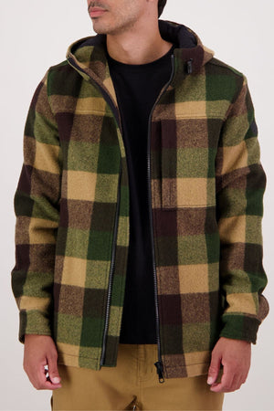 A person in a green, brown, and tan plaid Swanndri Hudson Hoodie made from New Zealand wool, layered over a black shirt and tan pants, stands against a plain light background with their face partially out of frame.
