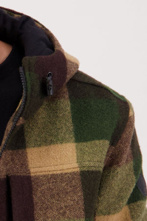Close-up of a person wearing a Swanndri Hudson Hoodie with green, brown, and tan plaid. This warm, hard-wearing jacket features a zipper and zippered chest pocket, with part of a black hoodie visible underneath.
